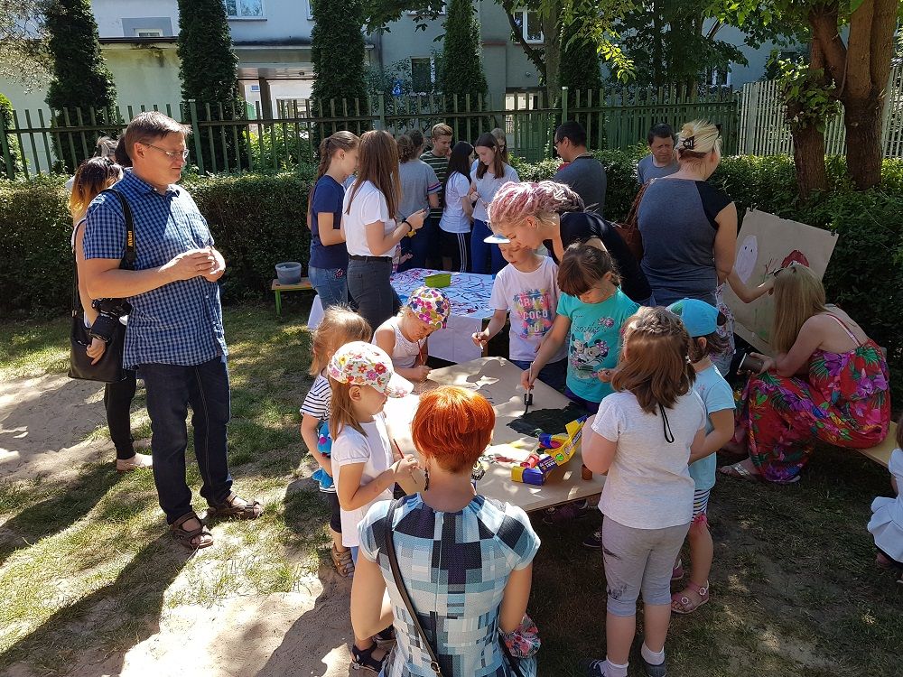 Dzieci na dworze zgromadzone przy stolikach, na których malują prace farbami
